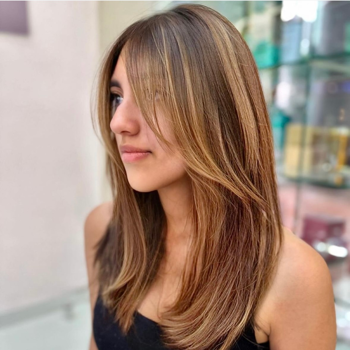 Primer plano de una persona con cabello largo con mechas, vestida con una blusa negra, mirando hacia un lado en una peluquería.