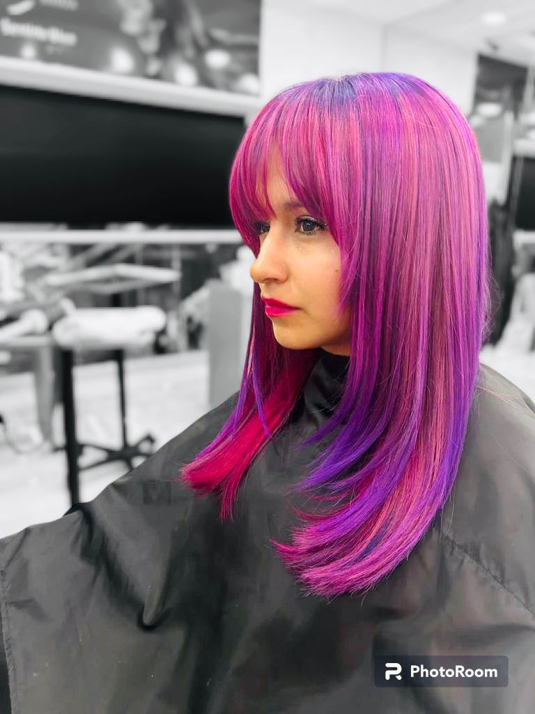 Mujer con cabello largo color magenta y flequillo, sentada con una capa de peluquería, sobre un fondo blanco y negro borroso.