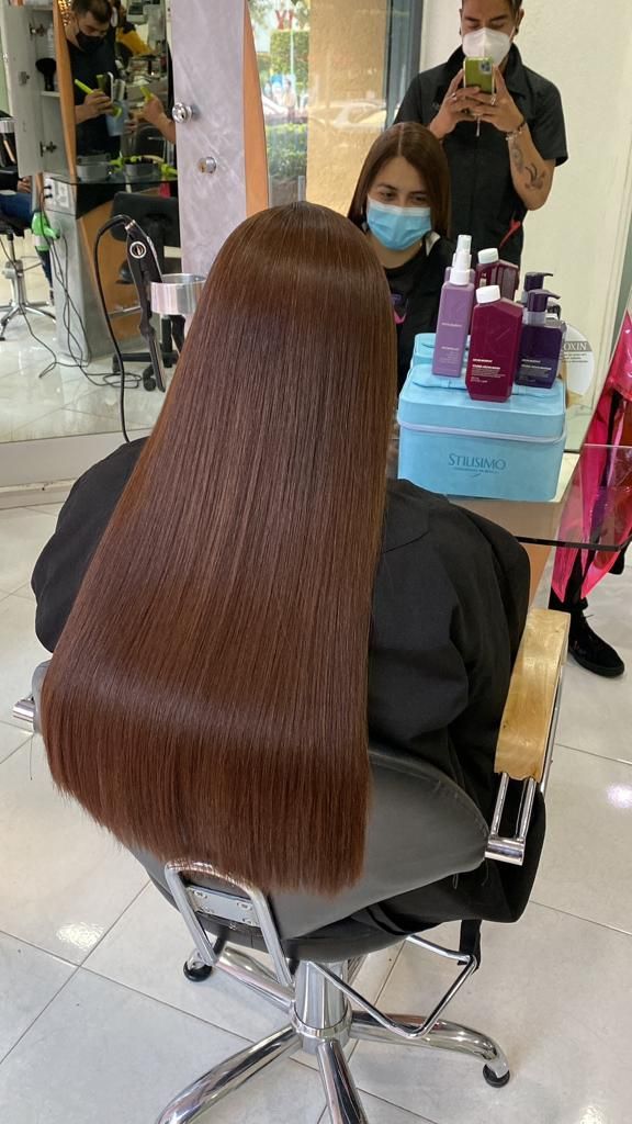 Clienta de salón con cabello largo, liso y castaño, sentada en una silla de peluquería, frente a un espejo con productos para el cabello cerca.
