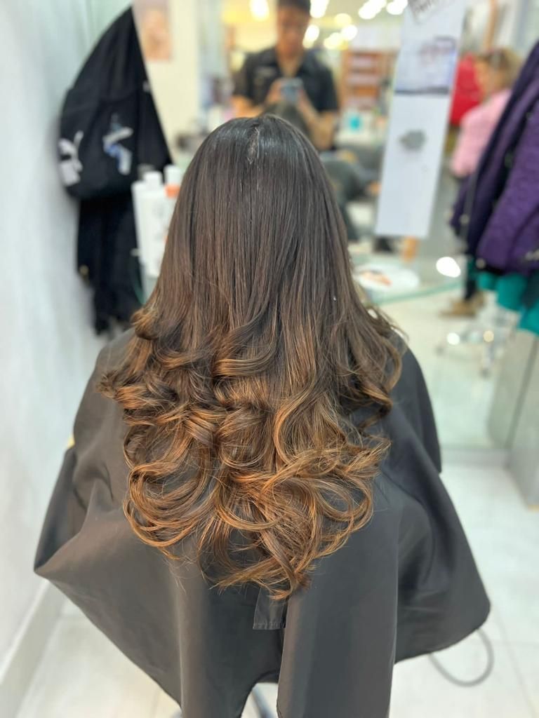 Vista posterior de una larga melena castaña ondulada con rizos más claros en una silla de peluquería.