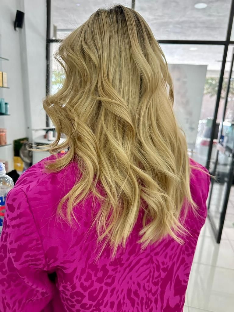 Vista trasera de una persona con cabello rubio ondulado que lleva una blusa rosa brillante en el pasillo de una peluquería.