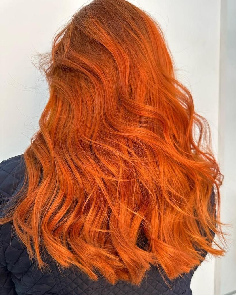 Primer plano de una melena ondulada de color naranja rojizo brillante que cae sobre una blusa oscura sobre un fondo claro.
