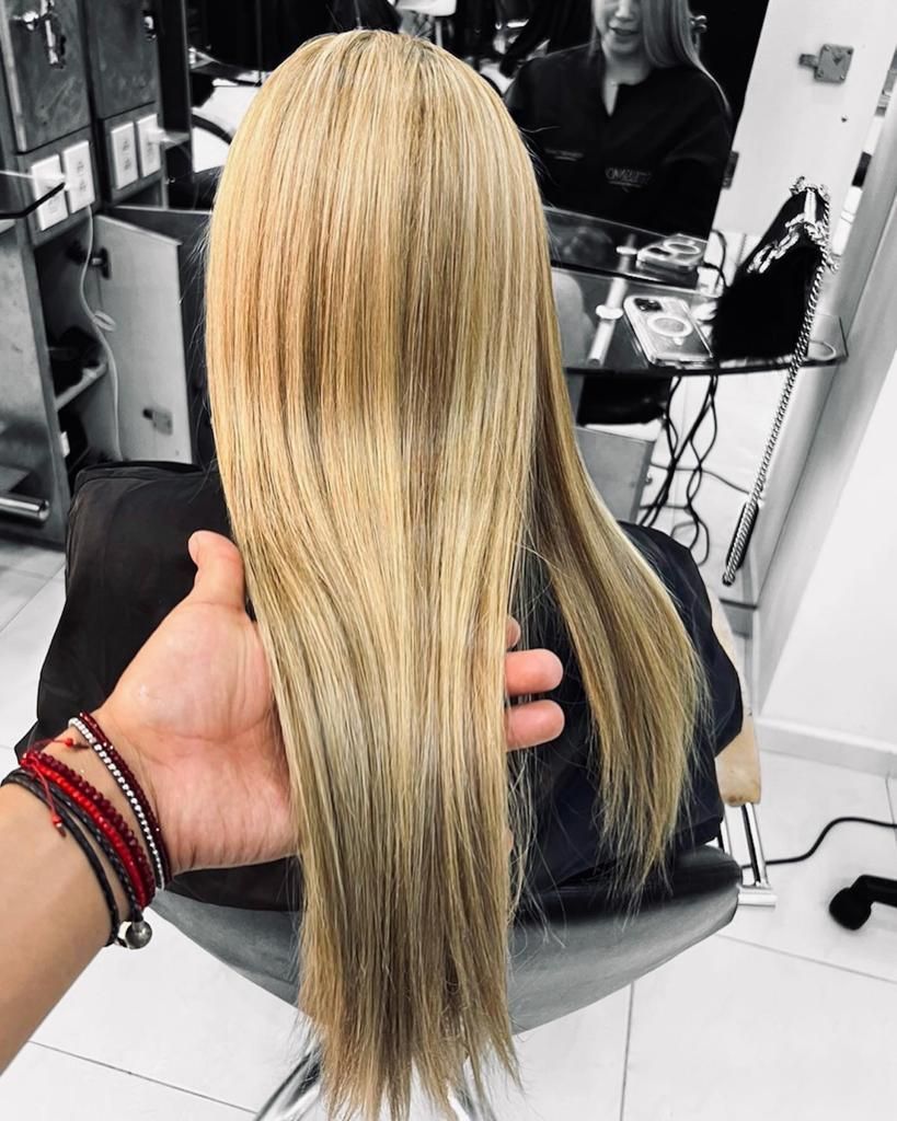Una mano sostiene una larga extensión de cabello rubio en una silla de peluquería, con un espejo y herramientas de peinado al fondo.