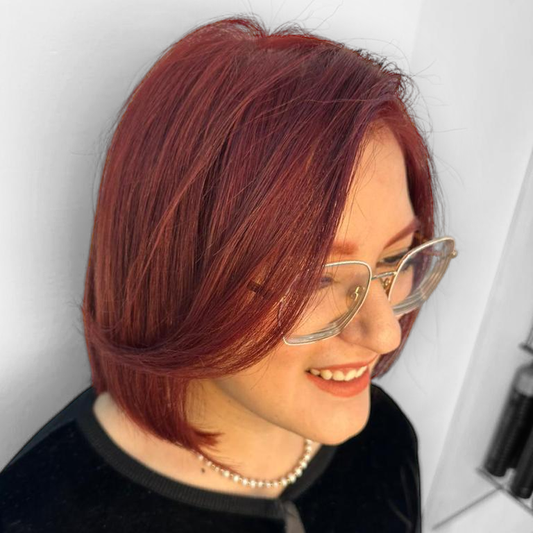 Persona con cabello corto y rojo y gafas sonriendo con una blusa negra sobre un fondo claro.