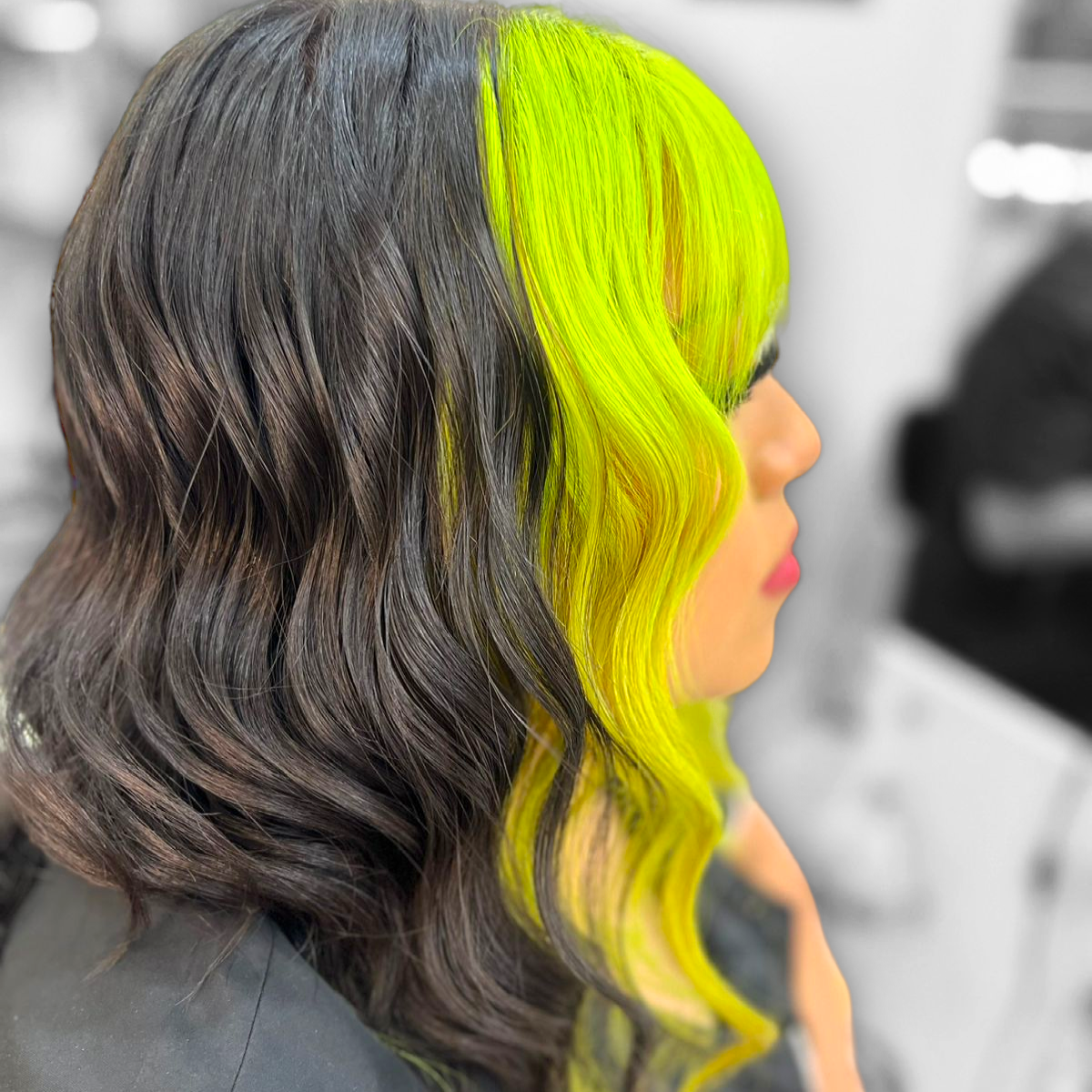 Vista lateral de cabello negro ondulado con reflejos frontales de color amarillo verdoso neón en un salón de belleza.