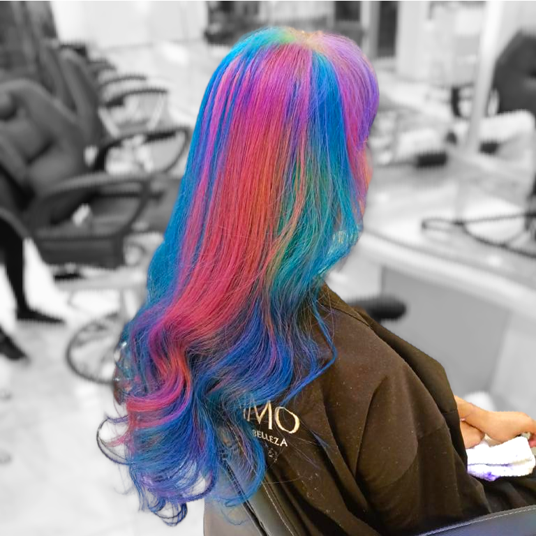 Persona sentada en una silla de peluquería con el pelo largo de colores del arcoíris, vista desde atrás.