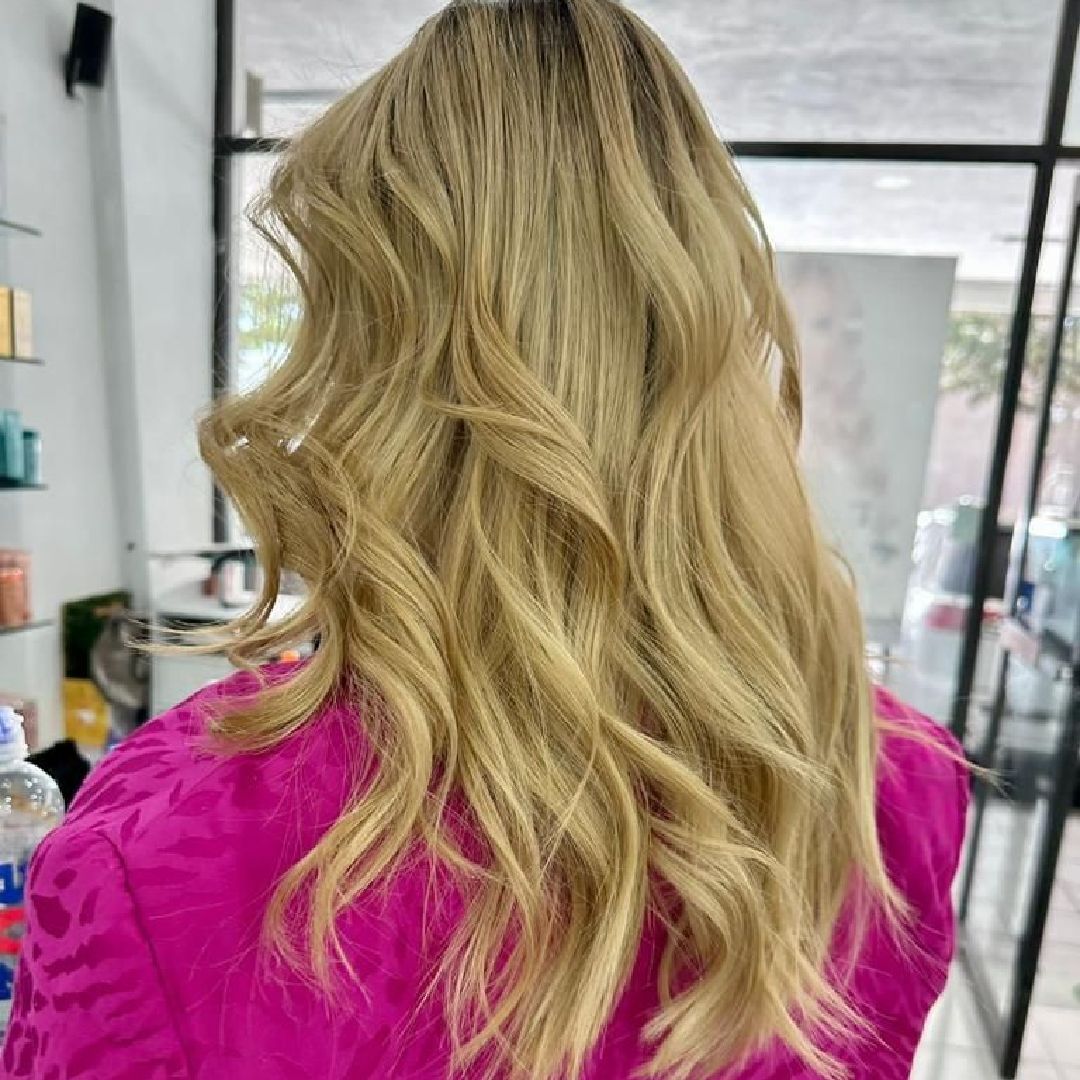 Cabello rubio ondulado visto desde atrás, con una blusa rosa brillante en un salón de belleza.