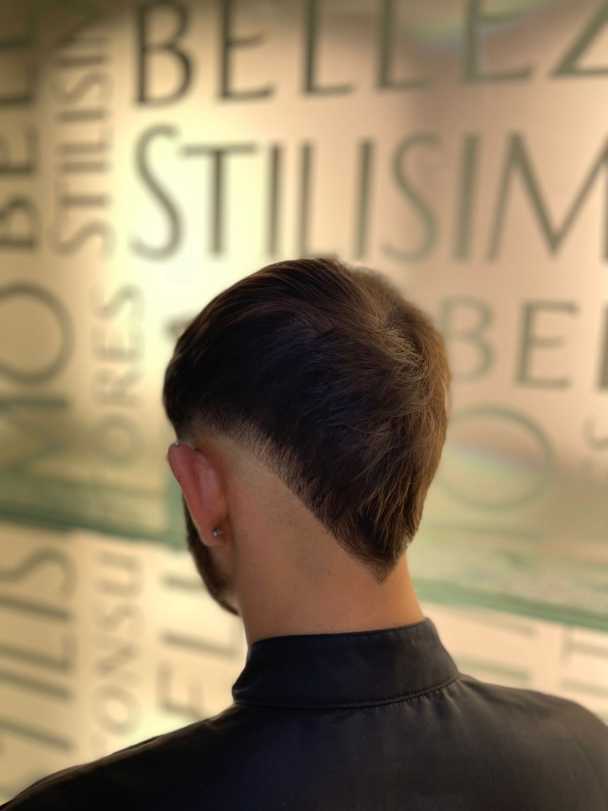 Persona con cabello corto y oscuro vista de espaldas, de pie frente a una pared tenuemente iluminada con grandes letras borrosas.