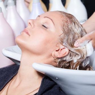 Una clienta está recostada en el lavabo de un salón de belleza mientras una estilista le enjuaga el cabello con una toalla blanca.