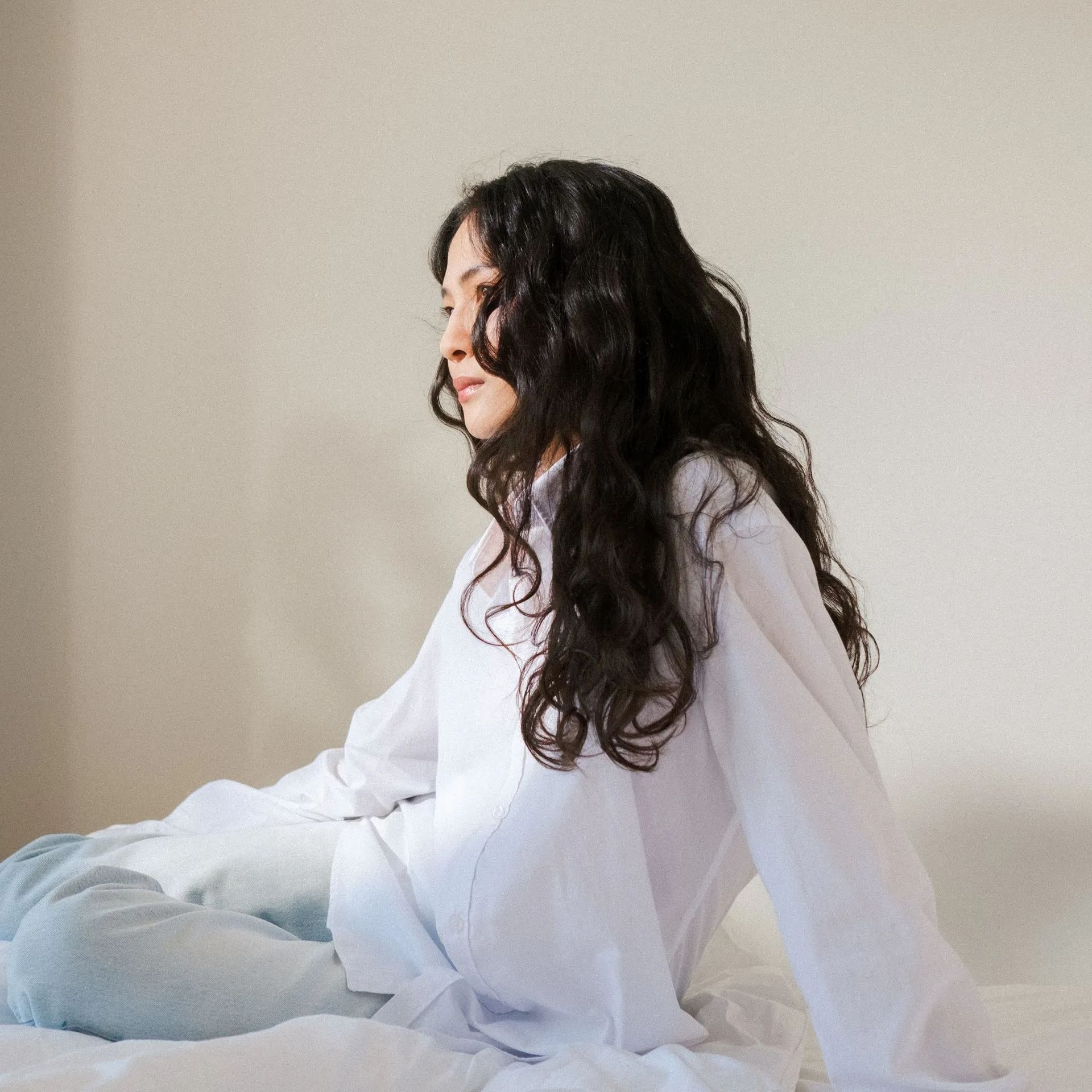 Mujer con camisa blanca sentada en una cama con sábanas azul claro, mirando hacia un lado.