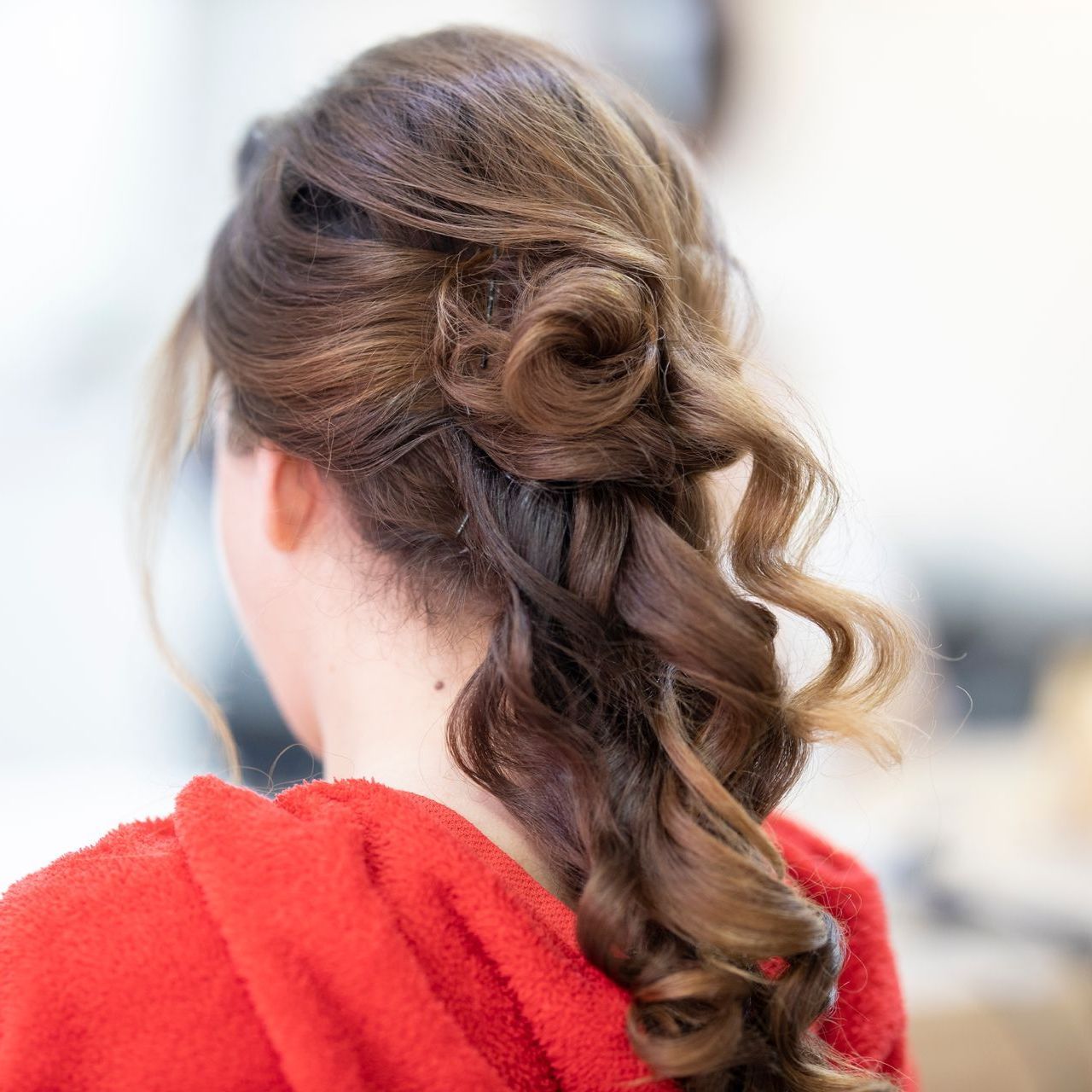 Mujer con cabello castaño rizado recogido en un moño, vestida con una túnica roja, vista desde atrás.
