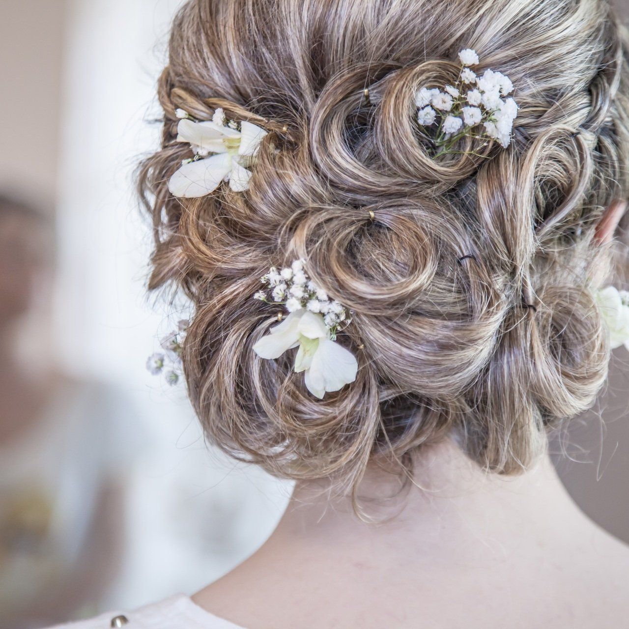 Intrincado recogido rubio con accesorios florales blancos para el cabello, visto desde atrás con iluminación suave.