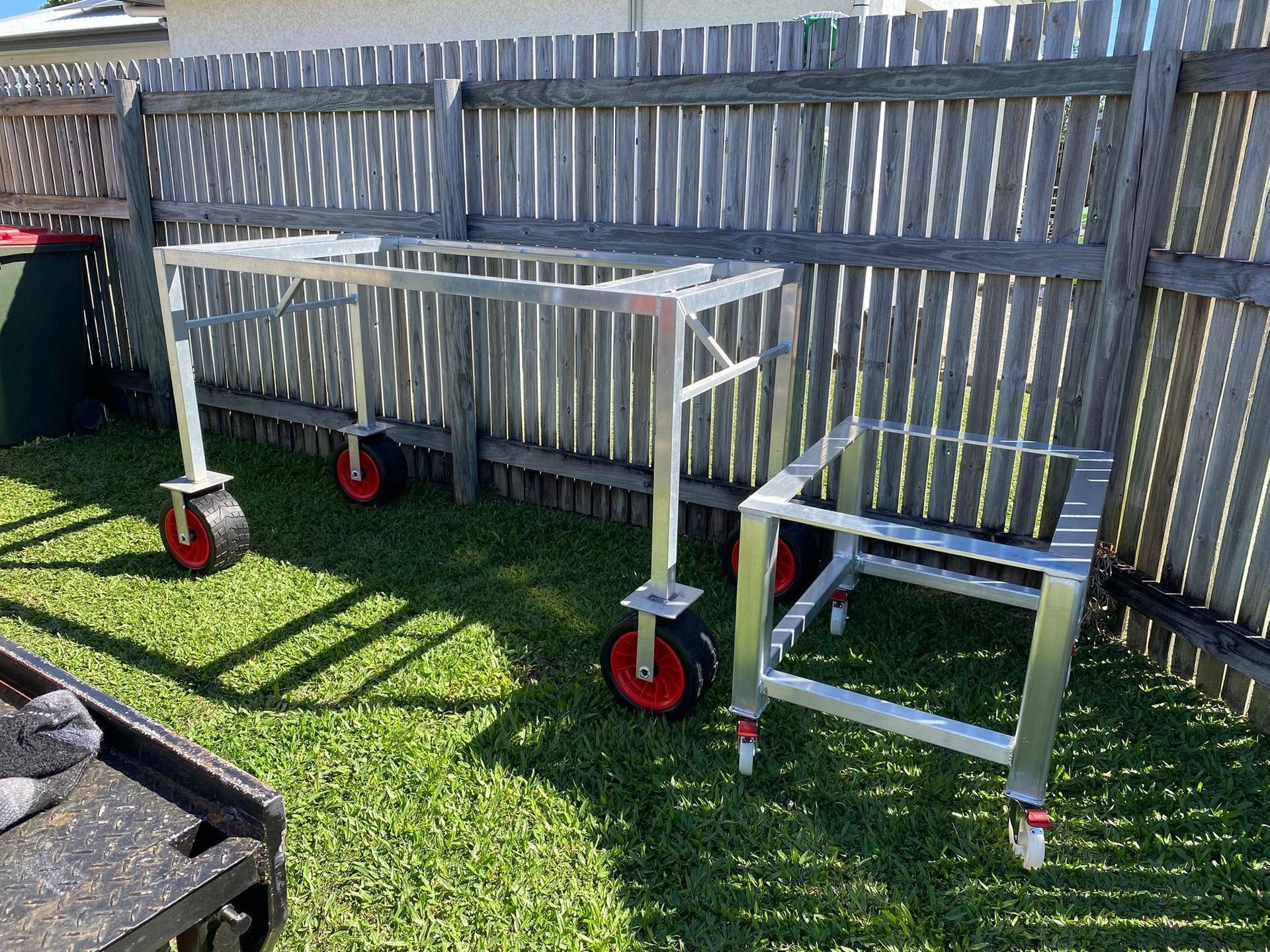 Two aluminum metal frames on wheels stand on a grassy lawn in front of a wooden fence.