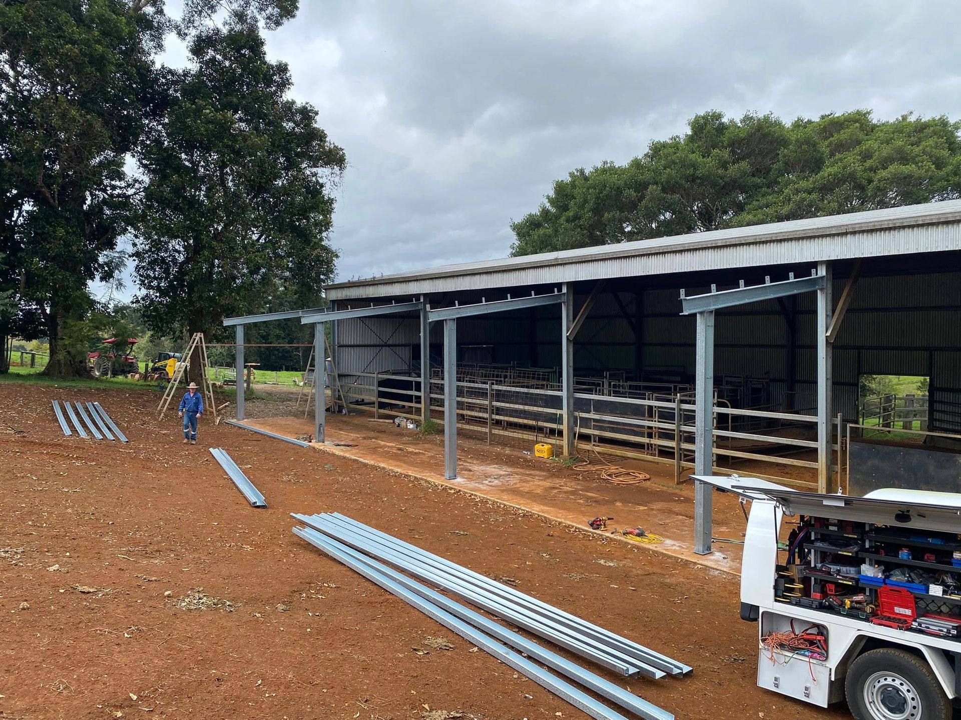 Construction workers build a metal-framed shed addition on a rural property with piles of steel beams in the dirt yard.