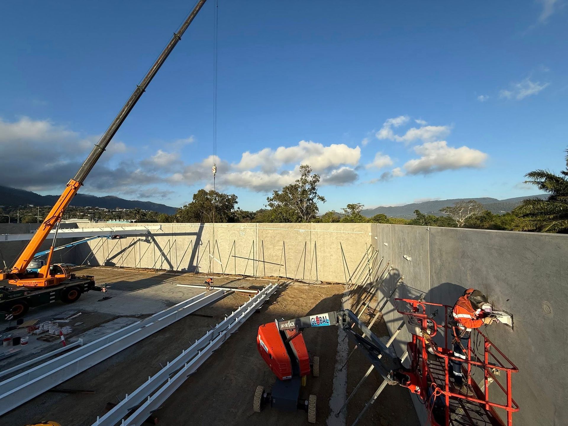 A crane and a worker in an aerial lift work on a large concrete wall at a construction site under a clear blue sky.