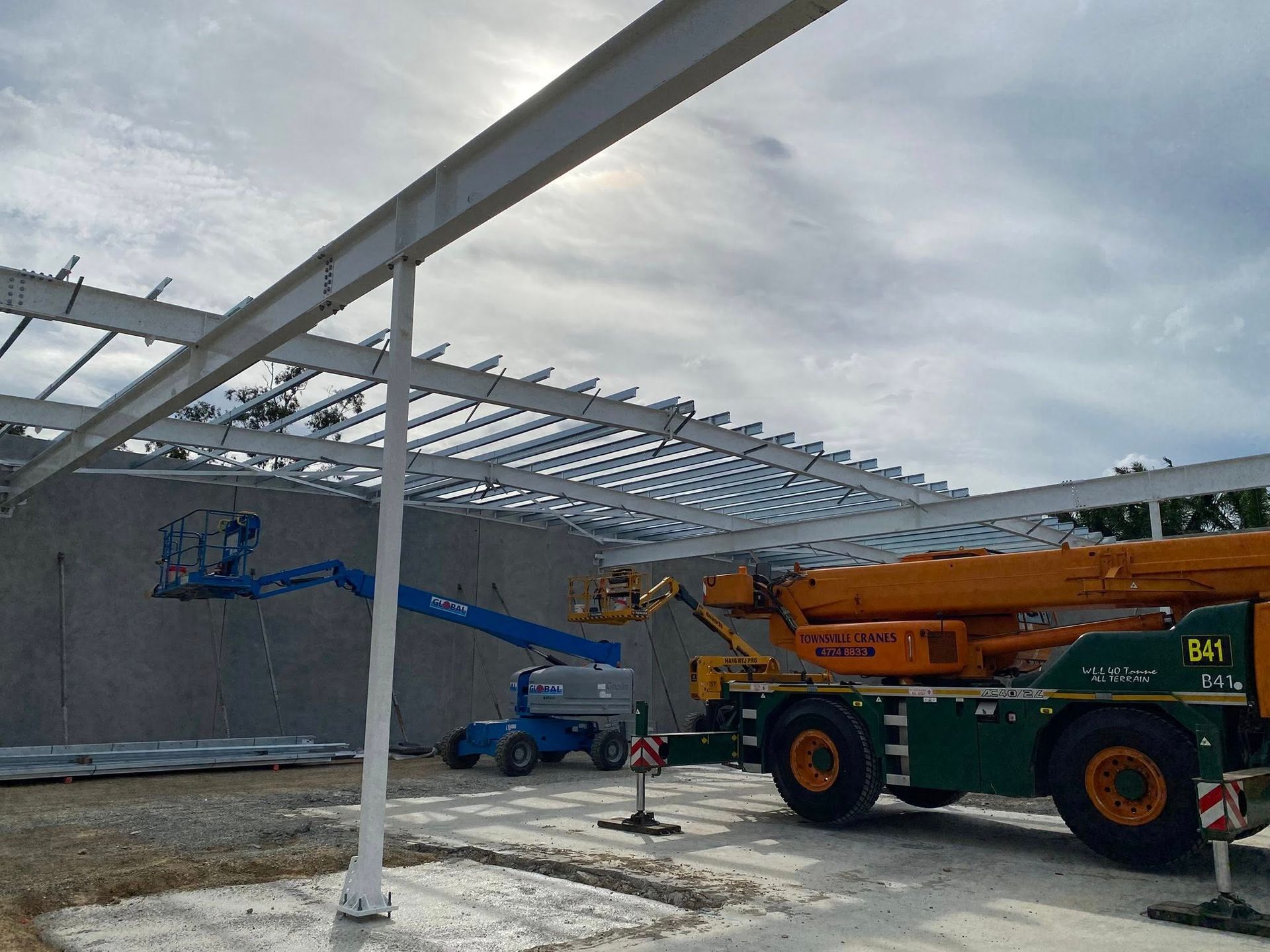 A construction site with a blue articulating boom lift and a yellow crane working on a steel building frame.