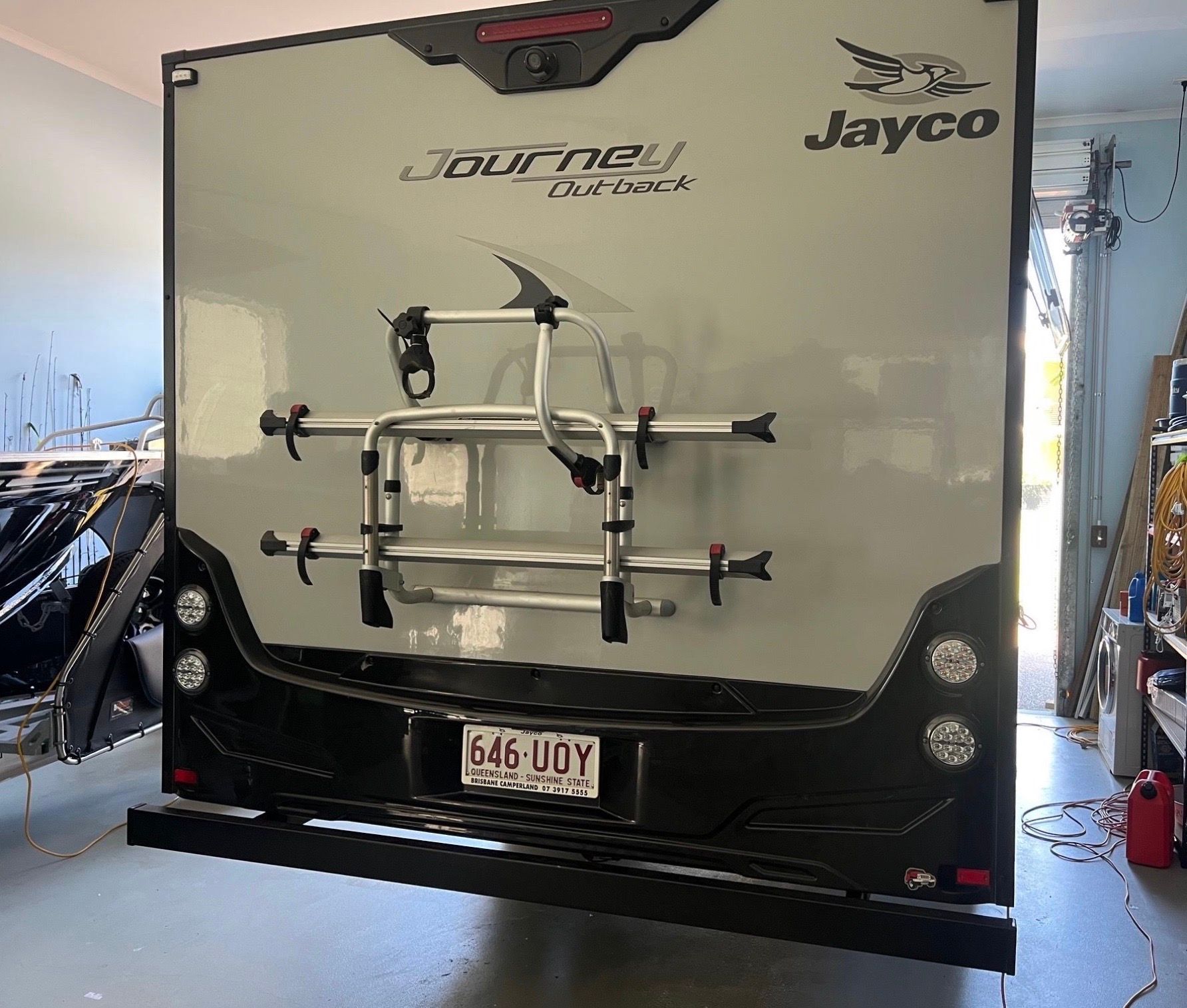 The rear exterior of a gray Jayco caravan parked indoors, featuring a mounted bicycle rack and license plate 646-UDY.