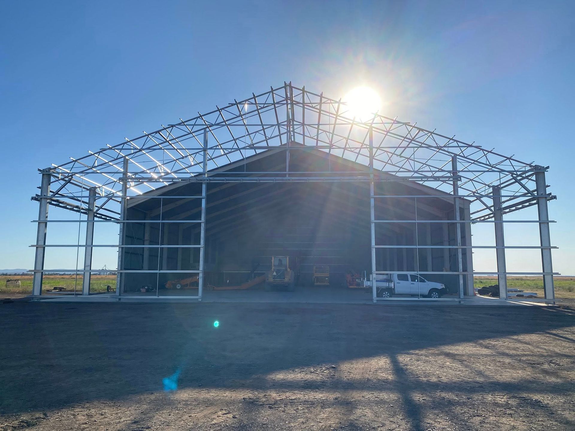 A large, incomplete metal warehouse frame stands on a gravel lot under a bright sun, with a white pickup truck inside.