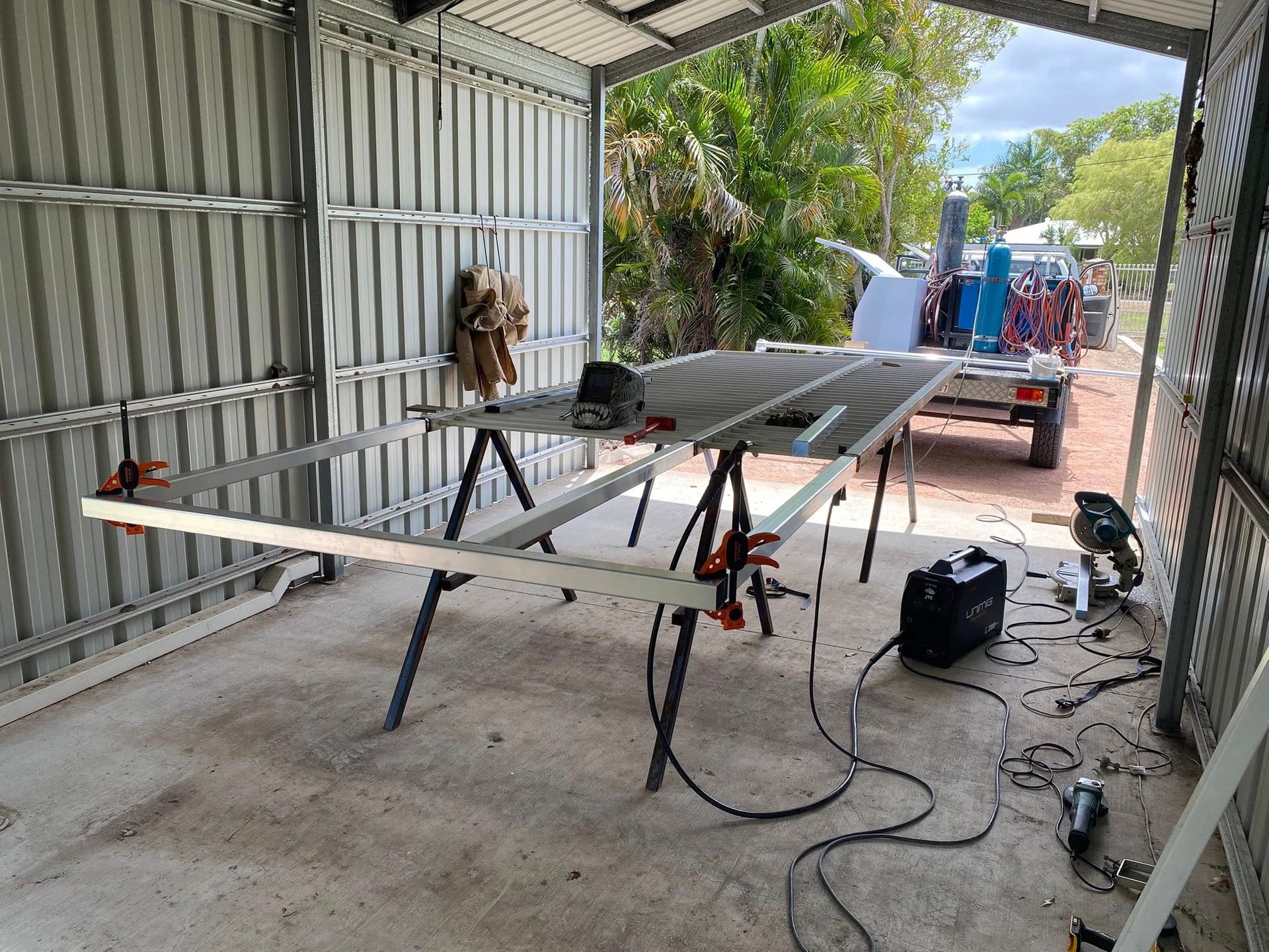 A metal frame sits on sawhorses inside a shed, with welding equipment nearby and a trailer visible through the opening.