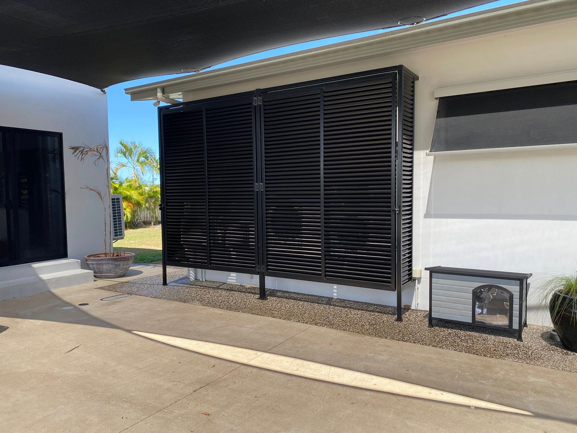 A black slatted privacy screen installed on the side of a white house next to a dog house on a gravel area.