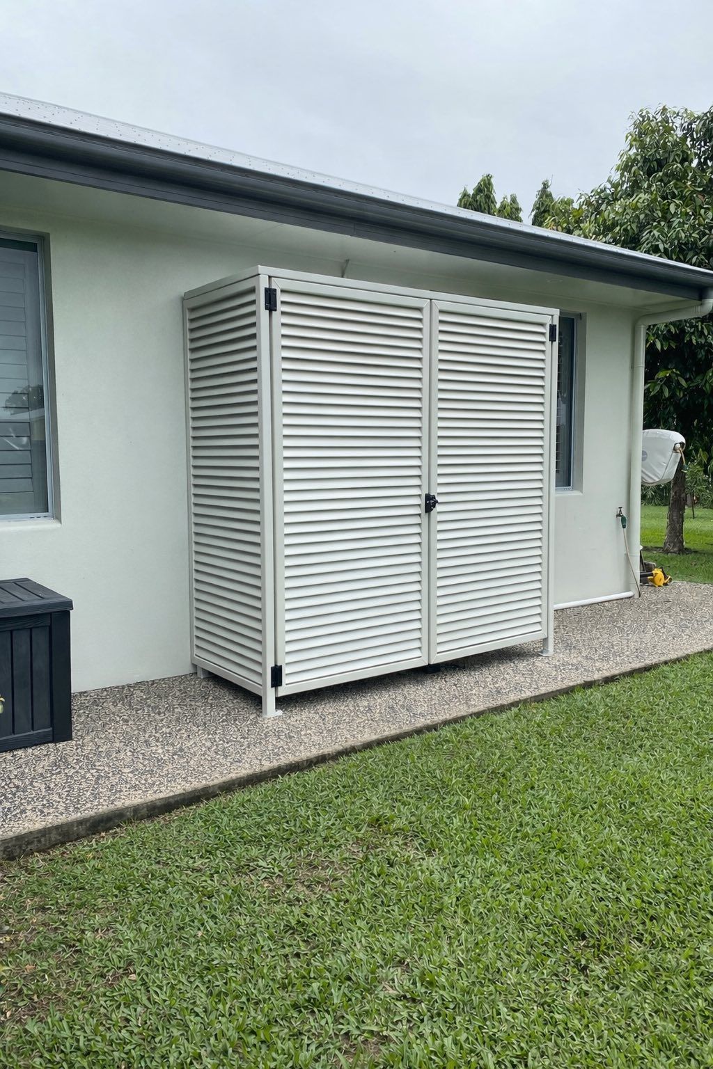 A white slatted storage cabinet stands against the exterior wall of a house on a gravel path next to a grass lawn.