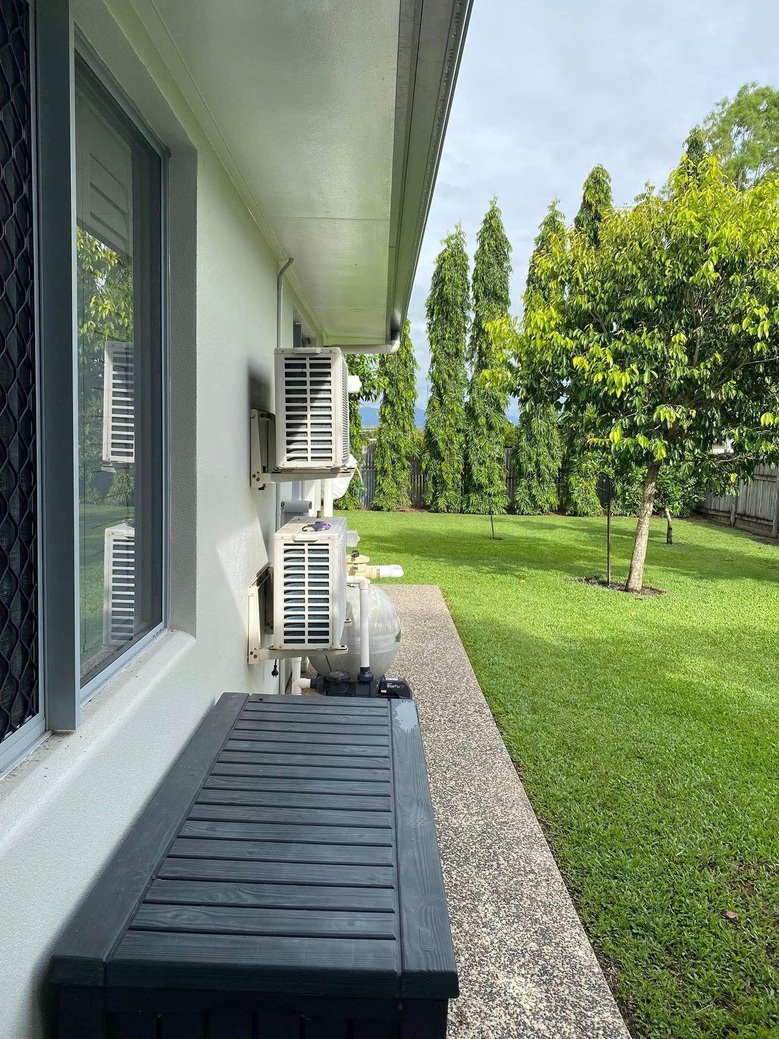 Two air conditioning units mounted on the side of a white building next to a gravel path and green lawn.