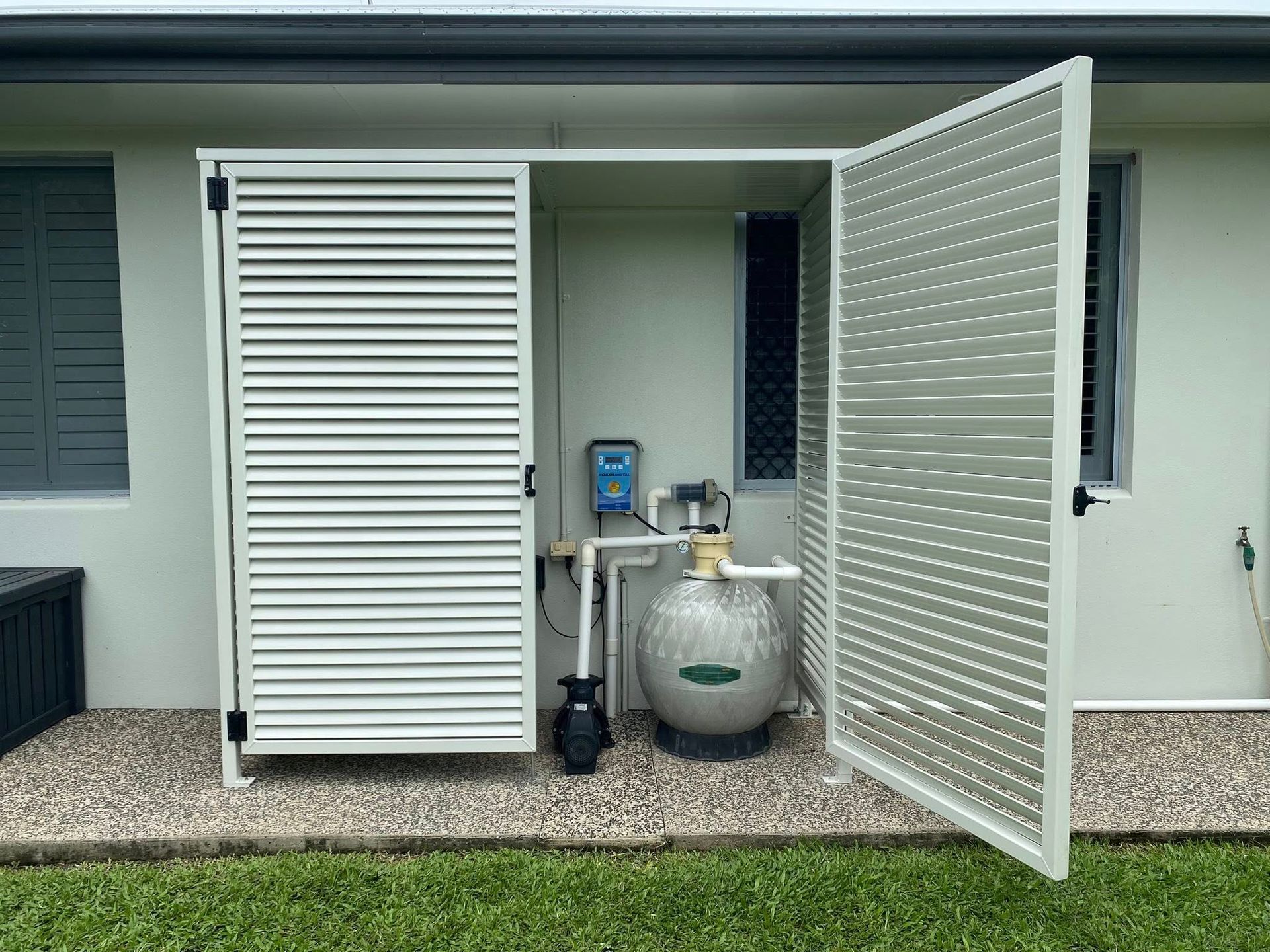 A white slatted enclosure housing pool equipment, including a pump and a spherical filter, against a house exterior.