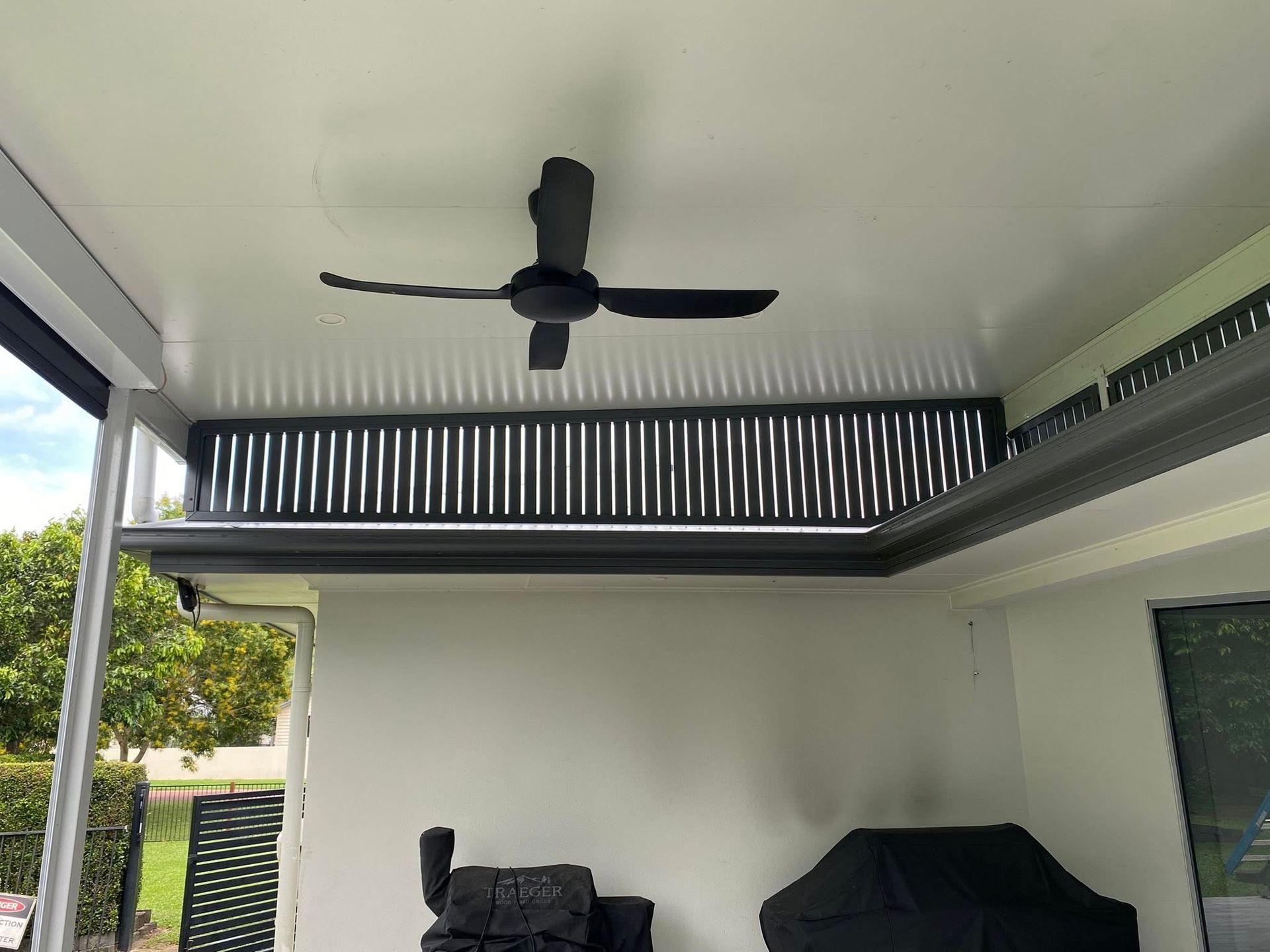 A covered outdoor patio with a black ceiling fan, vertical black privacy slats, and two black-covered items underneath.