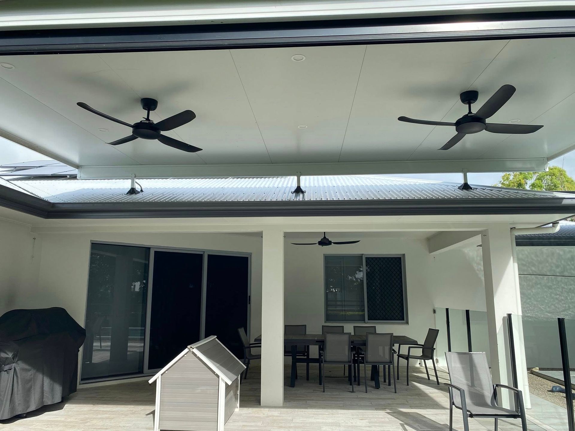 A covered outdoor patio with a dining set, a grill, a pet house, and two ceiling fans installed on the white ceiling.