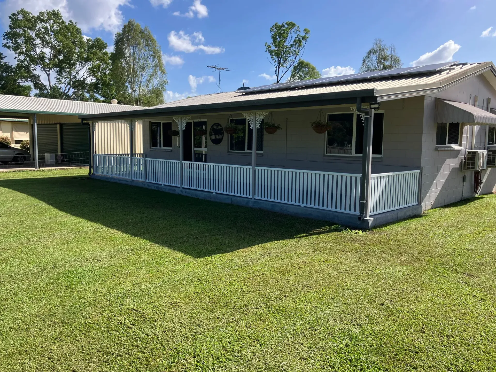 A light gray, single-story house with a white wraparound porch, set on a bright green lawn under a sunny blue sky.