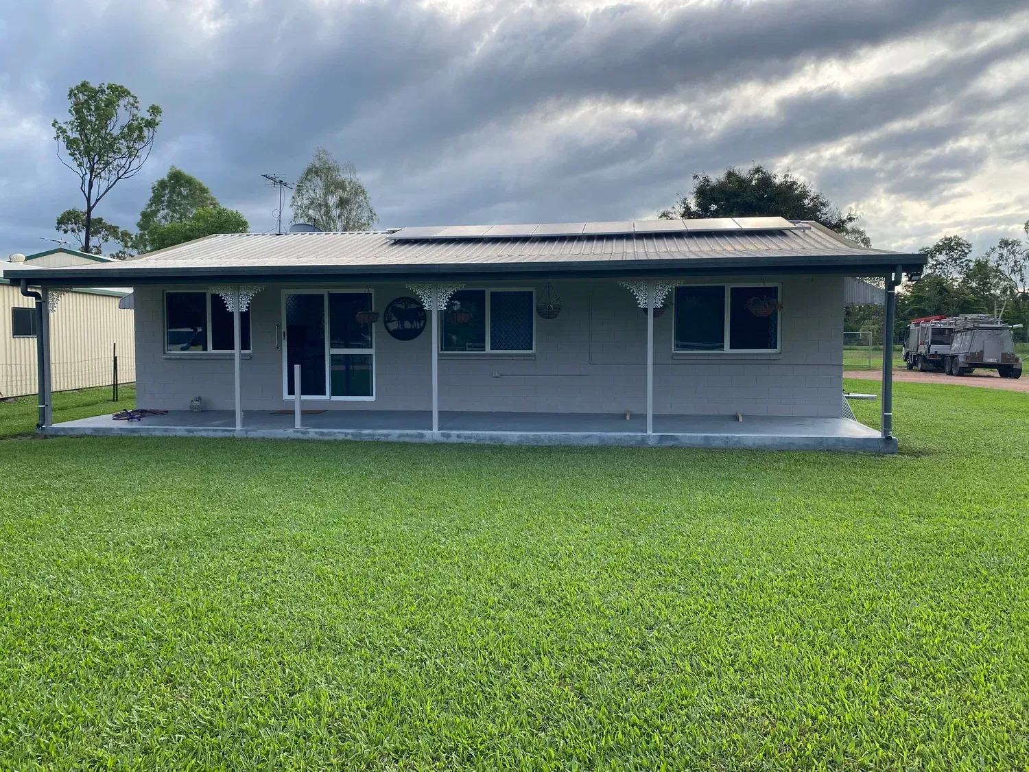 A single-story grey house with a front porch, a metal roof with solar panels, and a large lawn under a cloudy sky.