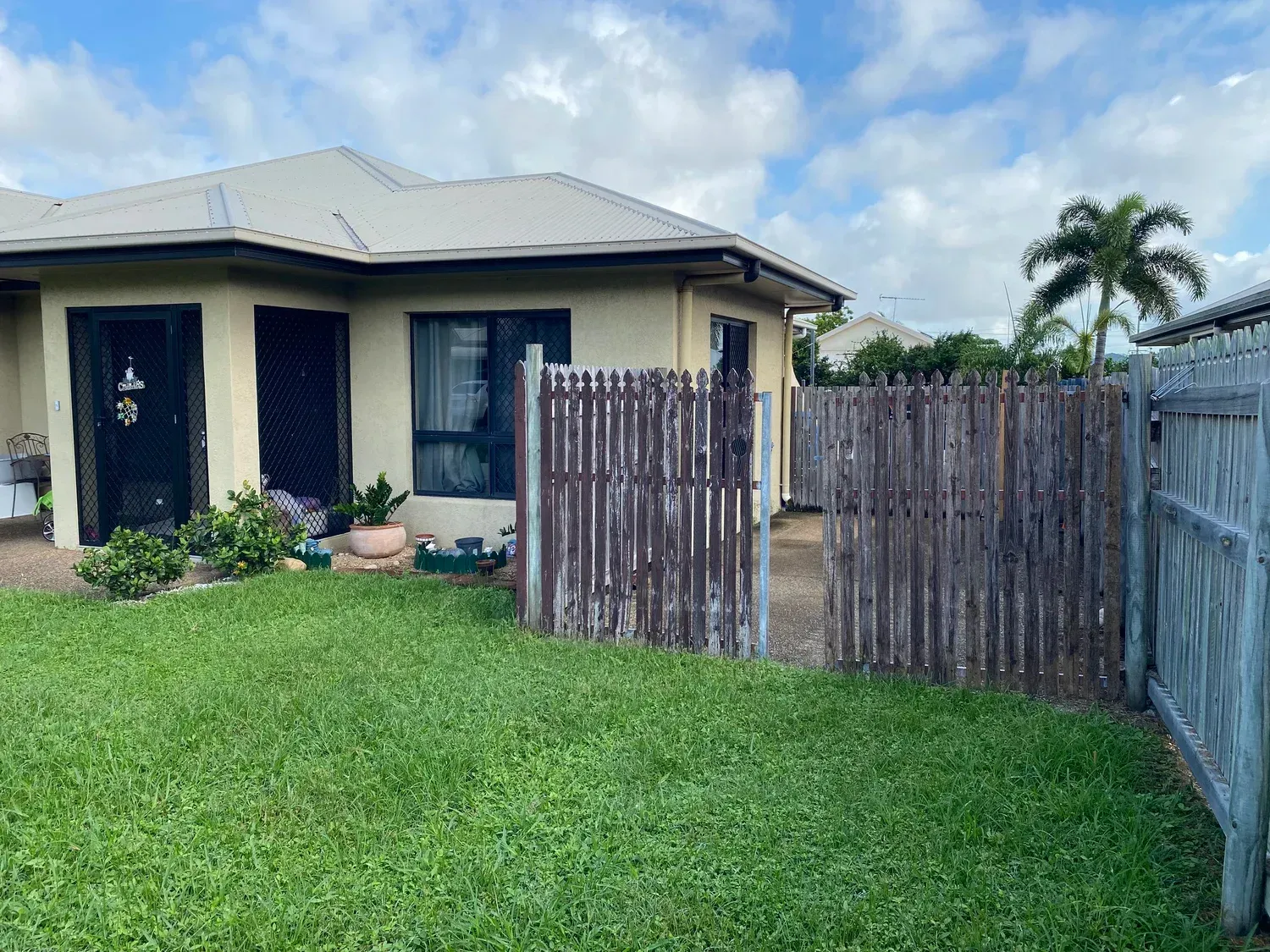 A single-story tan house with a gray roof, black doors, and a weathered wooden fence in a grassy front yard.
