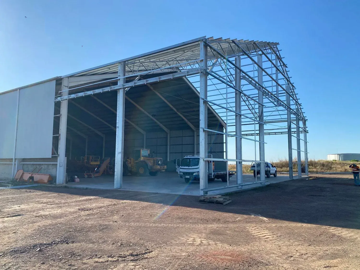 A partially constructed large metal shed frame stands on a dirt lot under a clear blue sky, with a truck parked inside.