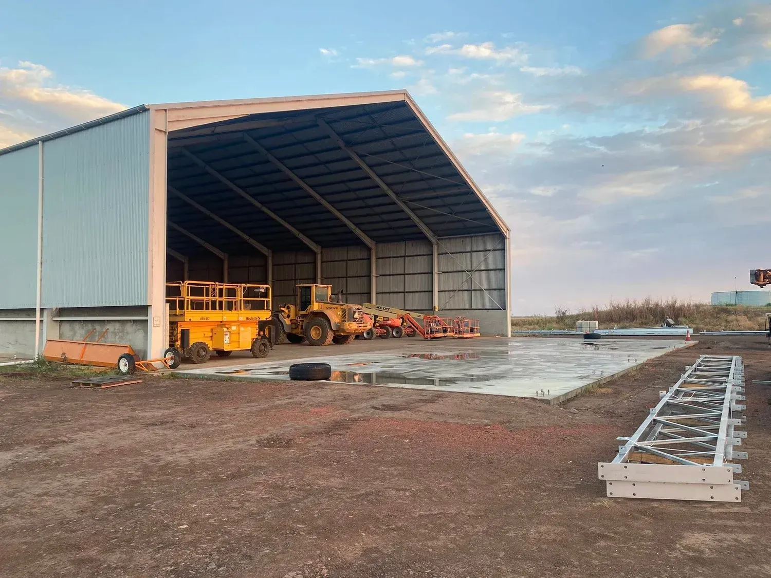 A large, open-sided metal equipment shed houses yellow machinery and construction items under a cloudy sky.
