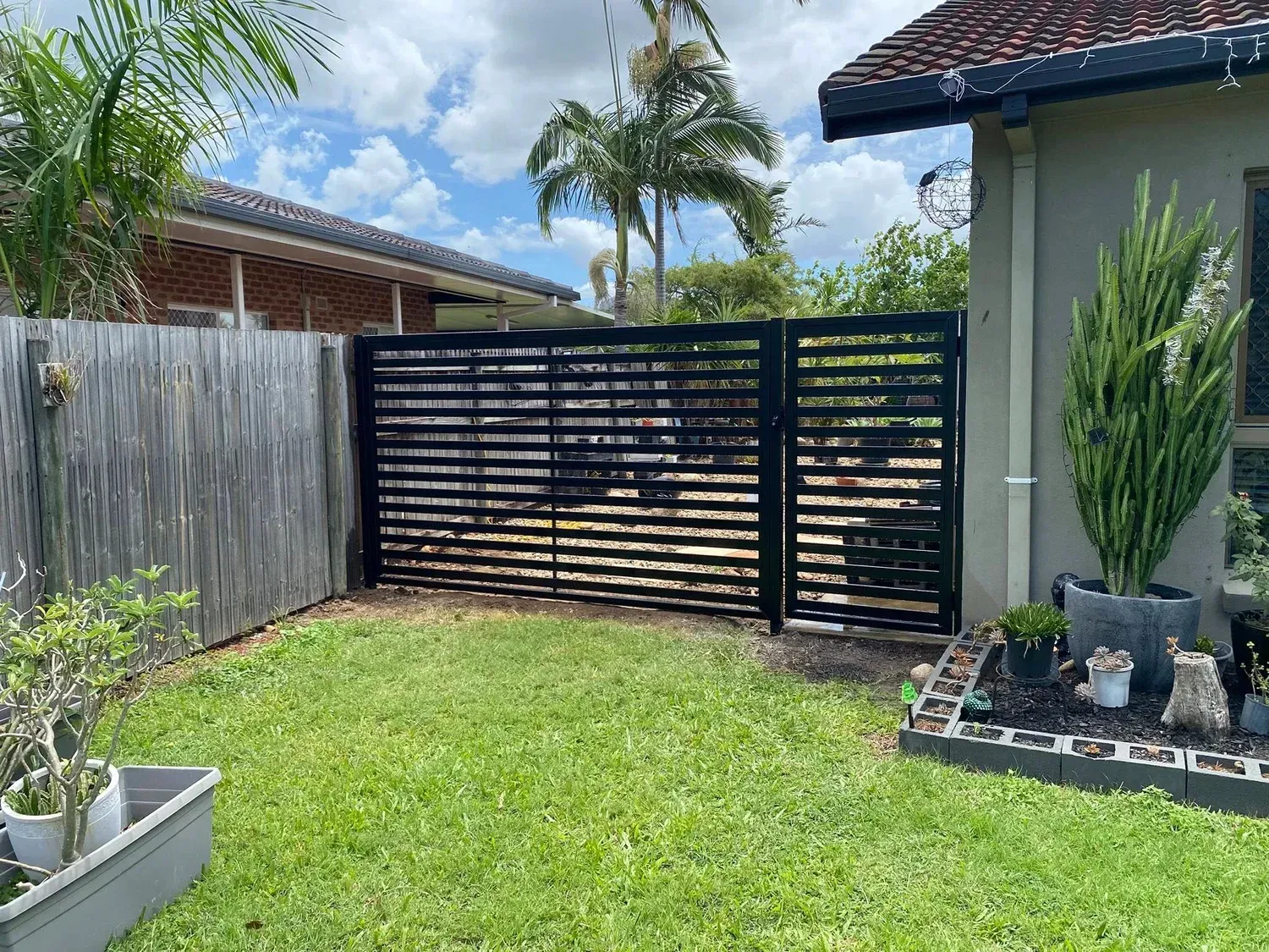 A black horizontal-slat gate stands between a wooden fence and a house, with a grass lawn and potted plants in the yard.