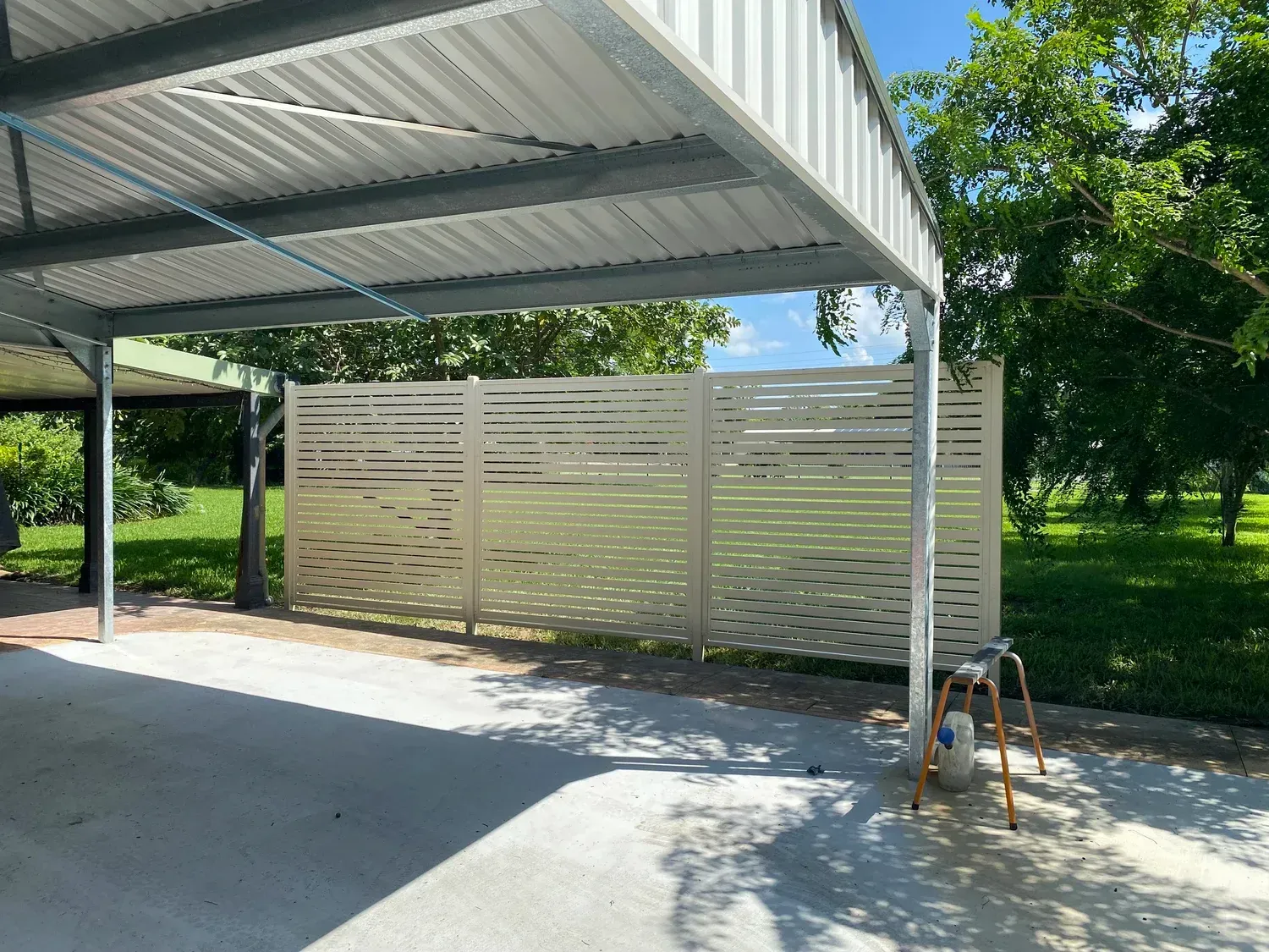A cream-colored, slatted privacy screen stands under a metal carport next to a grassy yard on a sunny day.