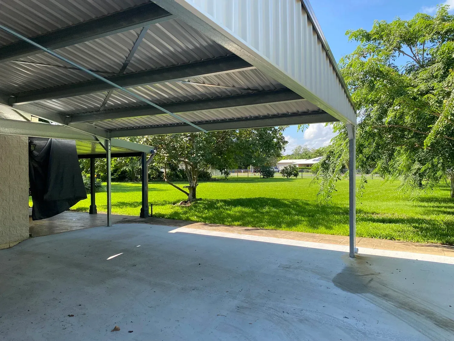 A view from under a metal carport looking out toward a sunny, grassy backyard with trees.