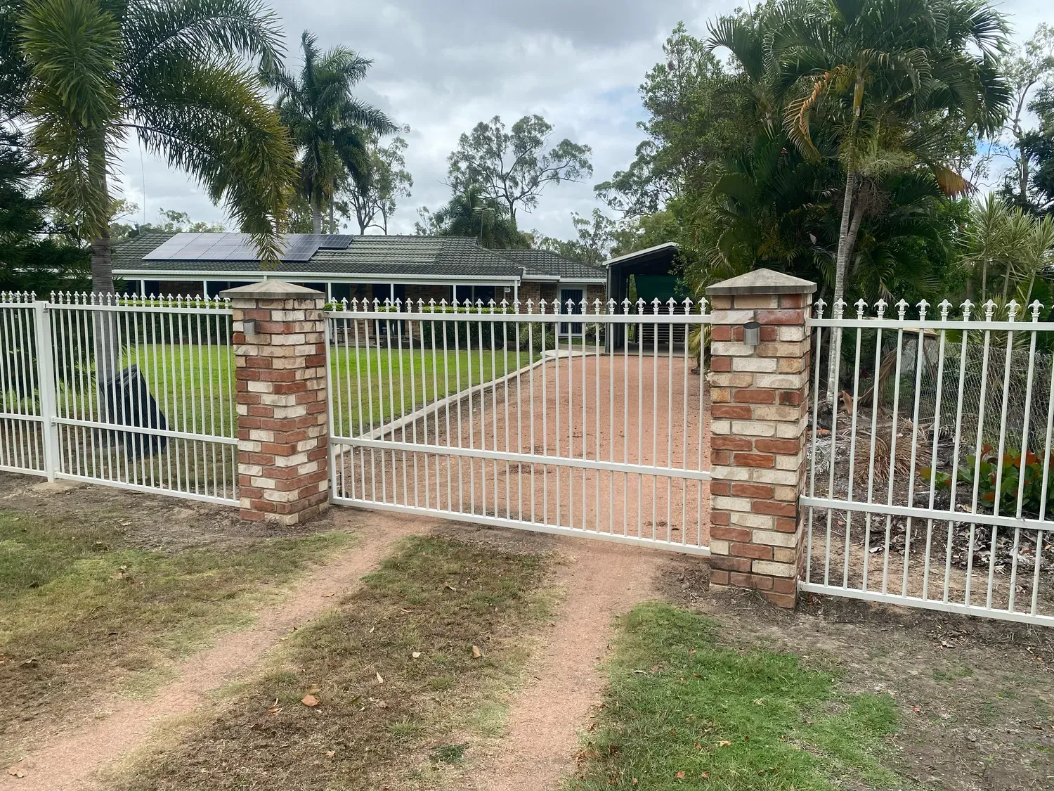 A white metal driveway gate with brick columns stands in front of a single-story house with a lawn and solar panels.