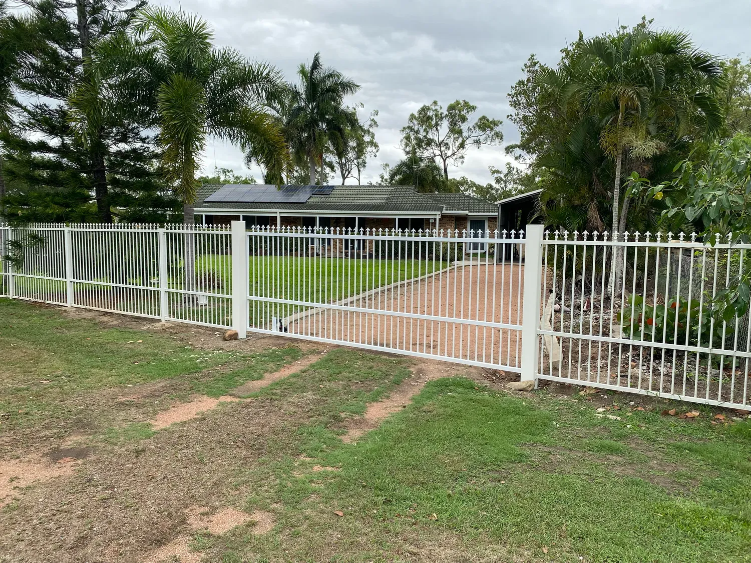 A white metal fence with a wide gate fronting a house, a paved driveway, and palm trees under a cloudy sky.