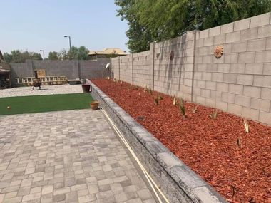 Backyard with paver patio, artificial turf, and a planter box with red mulch and plants against a cinder block wall.
