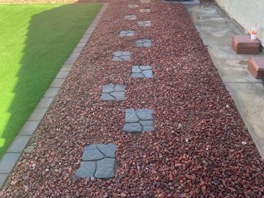 A garden path with square stepping stones through red gravel, bordered by green grass and concrete.