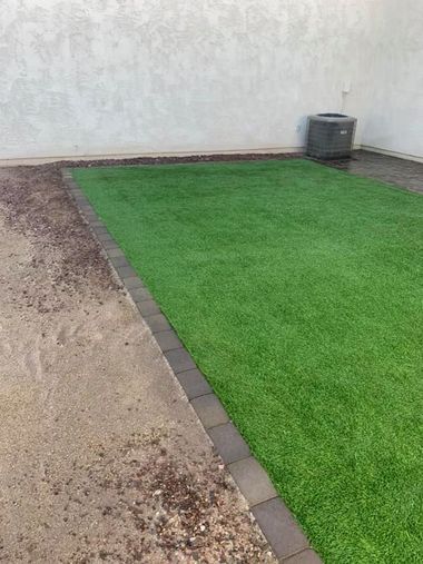 Green artificial turf bordered by dark pavers, next to a concrete surface, with a wall and AC unit in the background.