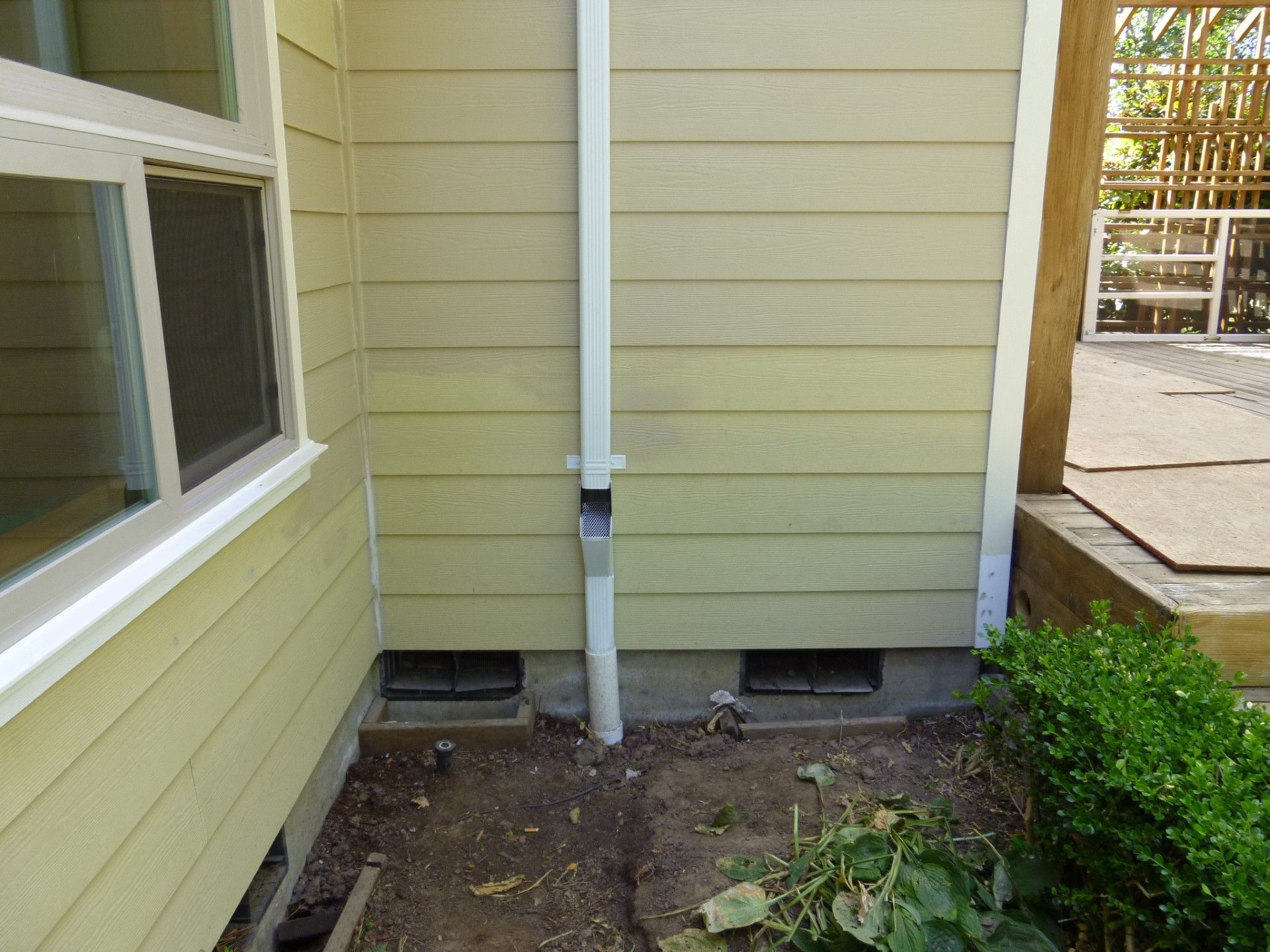 Side yard between yellow siding walls with a white downspout, gravel, and a concrete patio edge.