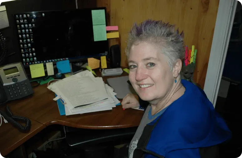 Smiling woman at a desk with a computer, papers, and colorful sticky notes in a wood-paneled room
