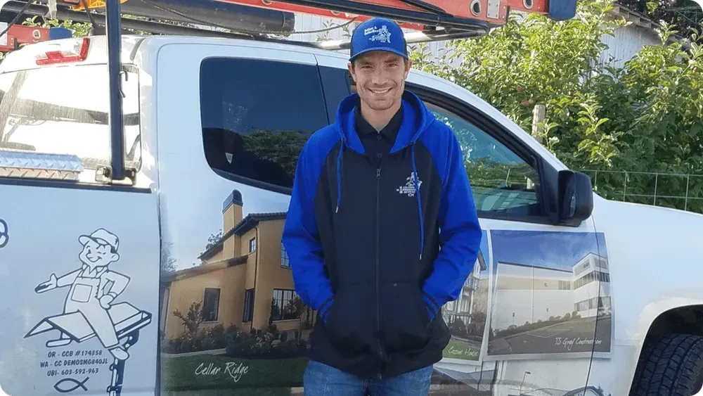 Smiling man in blue jacket and cap standing beside a white truck with a mural on the door