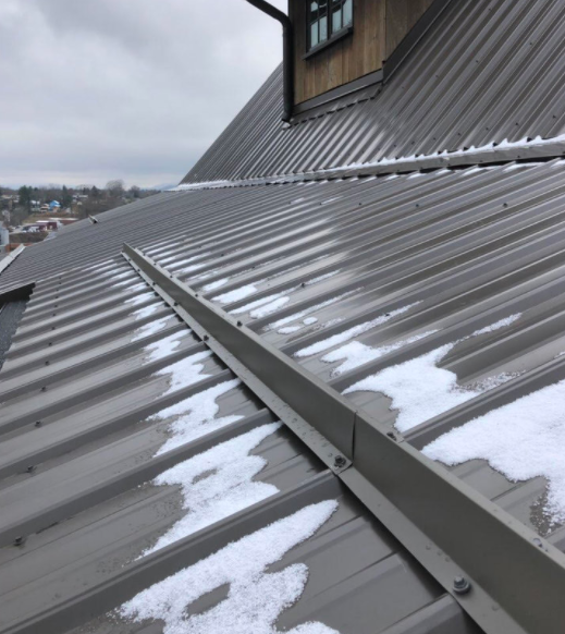 Close-up of a gray metal roof ridge with shingles and a wooden house roof in the background