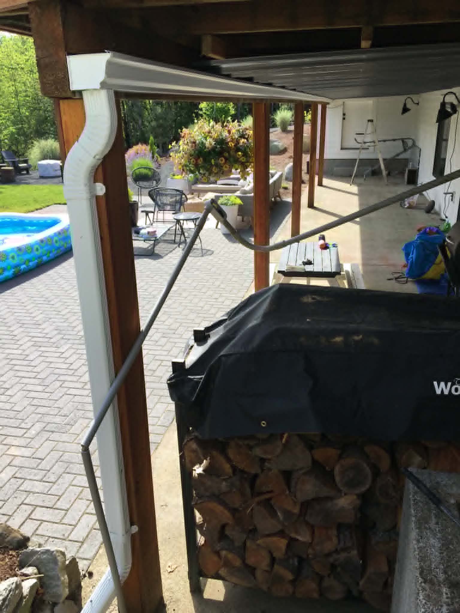 Covered backyard patio beside a pool, with stacked firewood and a folded metal frame under the deck.