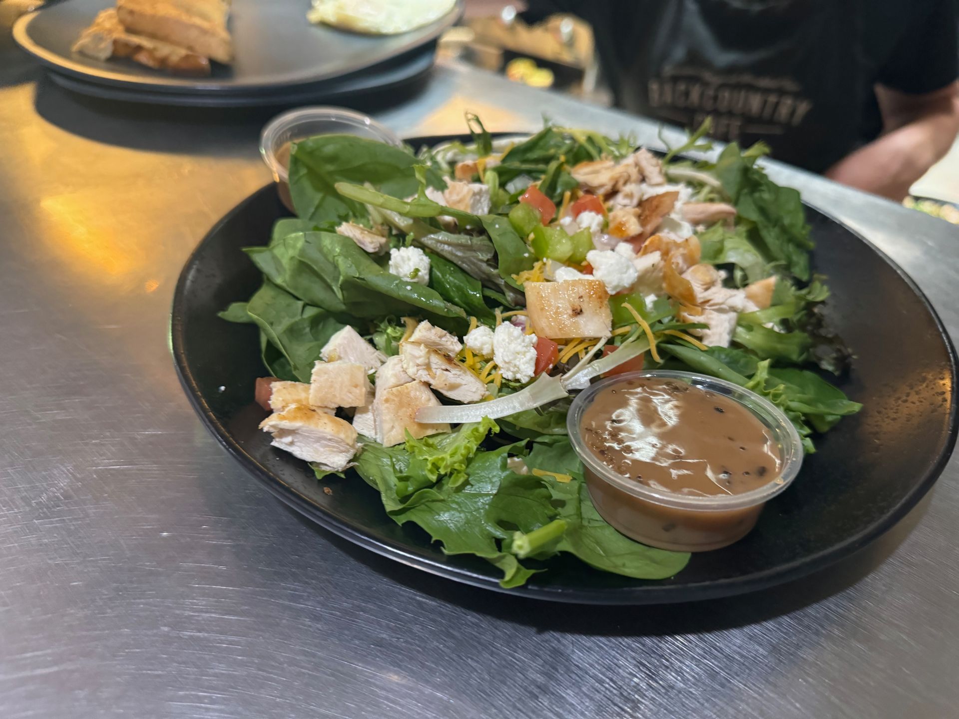 A salad with chicken and spinach on a black plate with a cup of dressing.