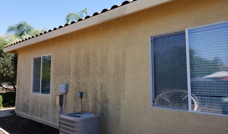 Exterior wall of a tan house with two windows, an air conditioner, and sloped roof under blue sky