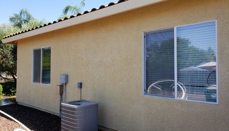 Tan stucco house exterior with two windows, a utility meter, and desert landscaping.